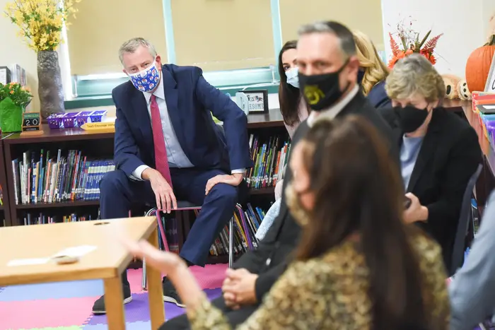 Mayor de Blasio sitting in a child sized chair in a classroom, in front of a shelf wtih children's books. Other adults are in the foreground